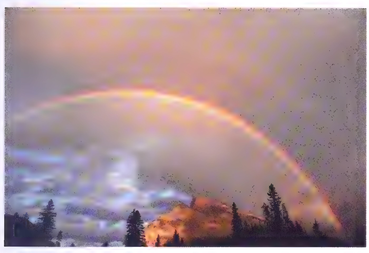 Photograph of a rainbow arching over a dark forest and a mountain, with the bow fading into the clouds at both ends.