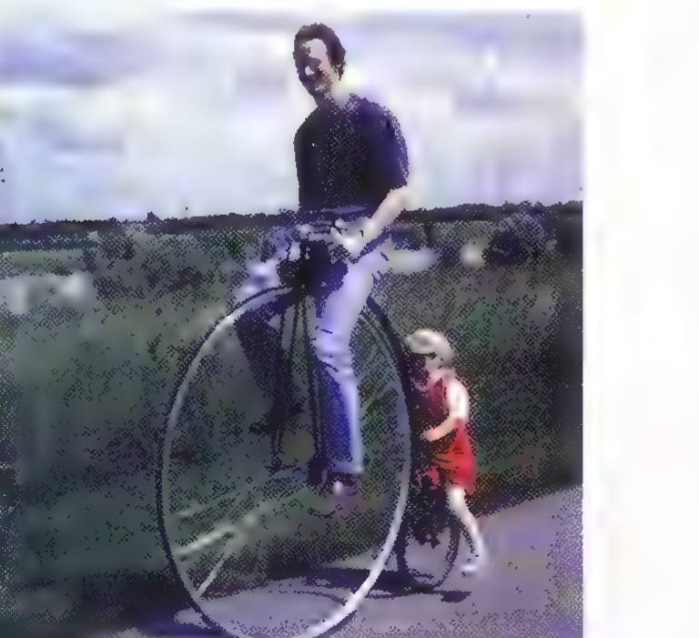 A man riding a penny-farthing bicycle with a very large front wheel and a small rear wheel, a child walking beside him.